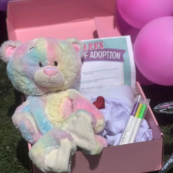 A colorful teddy bear sits in a Slime Party Box with an adoption certificate and pink balloons in the background.