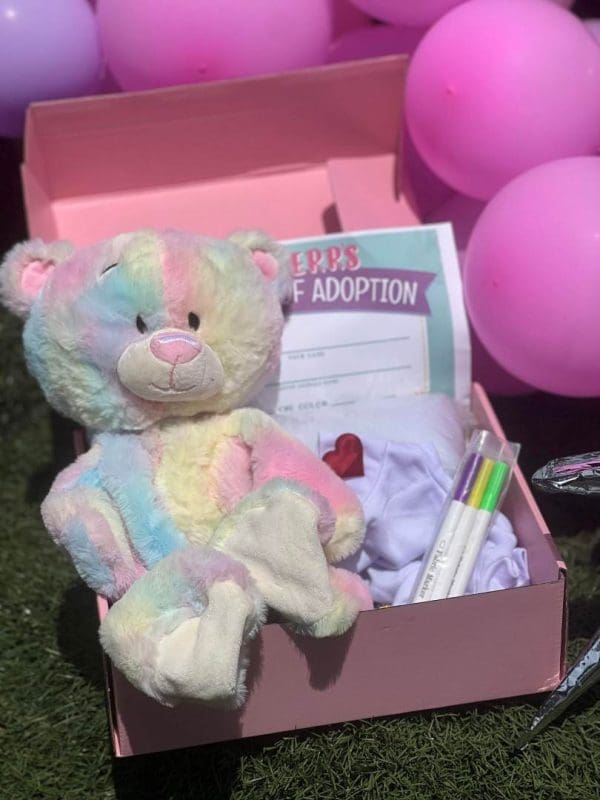 A colorful teddy bear sits in a Slime Party Box with an adoption certificate and pink balloons in the background.