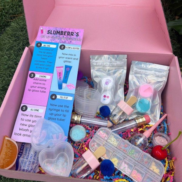 An open pink DIY Lip Gloss Party Box containing various tubes, glitter packets, mixing tools, and colorful decorations on a grass background.