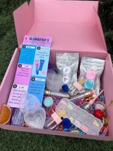 Open pink box delivered to a slumber party on grass, filled with various crafting supplies including glue, glitter, and colorful accessories.