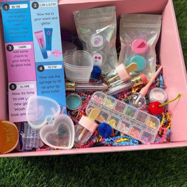 A colorful DIY Lip Gloss Party Kit with various components like glitter, scents, tubes, and a mixing guide spread out in a DIY Lip Gloss Party Box on grass.