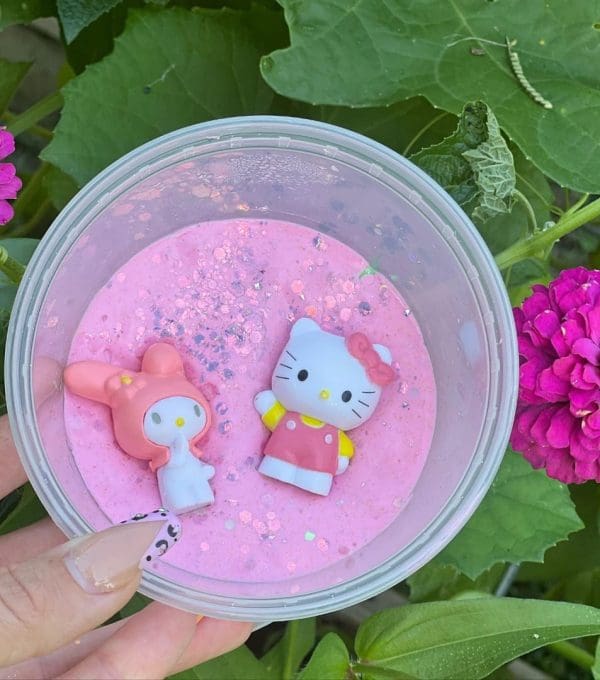 A hand holds a clear container of pink slime with glitter and two Hello Kitty figurines on top, surrounded by green leaves and pink flowers from the Enchanting Slime Boxes collection.