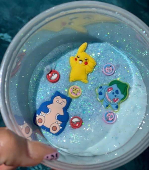 A hand holding a clear cup filled with enchanting slime and floating Pokémon-themed charms, including a Pikachu, with sparkly reflections visible.