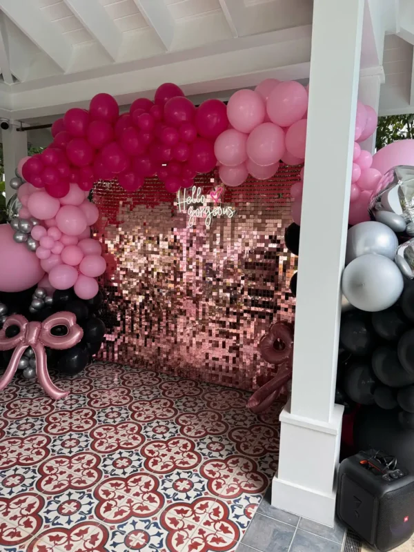 A Shimmer Backdrop Wall Rental with a "Hello Gorgeous" sign is framed by pink, black, and silver balloons, all set on patterned tile flooring under a covered area.