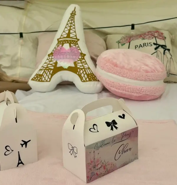 A "Welcome to Paris" gift box sits on a bed with Eiffel Tower and pink macaron pillows—perfect for photos when paired with the Shimmer Backdrop Wall Rental.