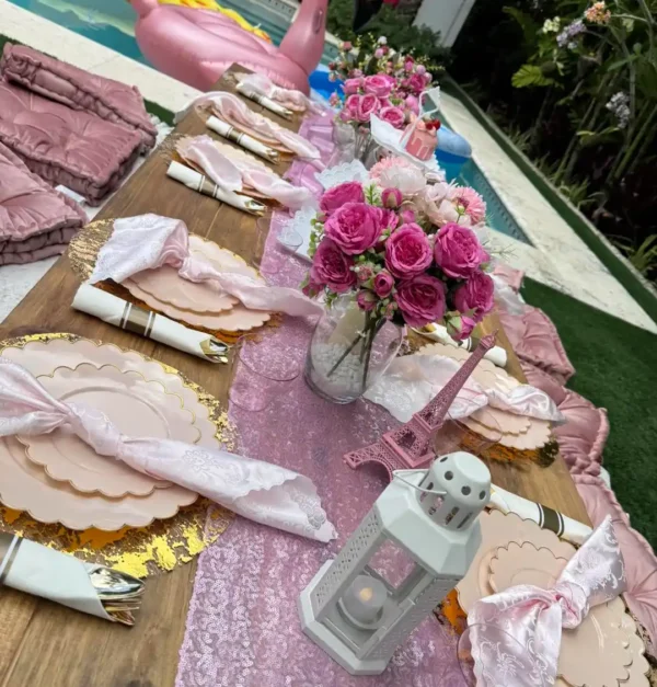 A wooden outdoor table set with pink and gold plates, floral centerpieces, a small Eiffel Tower, and a white lantern sits by the pool. An inflatable float and our Shimmer Backdrop Wall Rental add extra elegance to the scene.