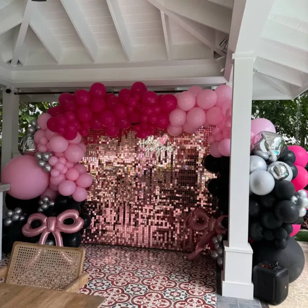 A pink and black balloon arch frames the Shimmer Backdrop Wall Rental under a white gazebo, with a wooden table and patterned tile floor in front.