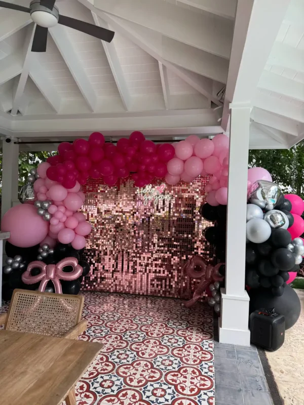 A pink and black balloon arch frames the Shimmer Backdrop Wall Rental under a white gazebo, with a wooden table and patterned tile floor in front.