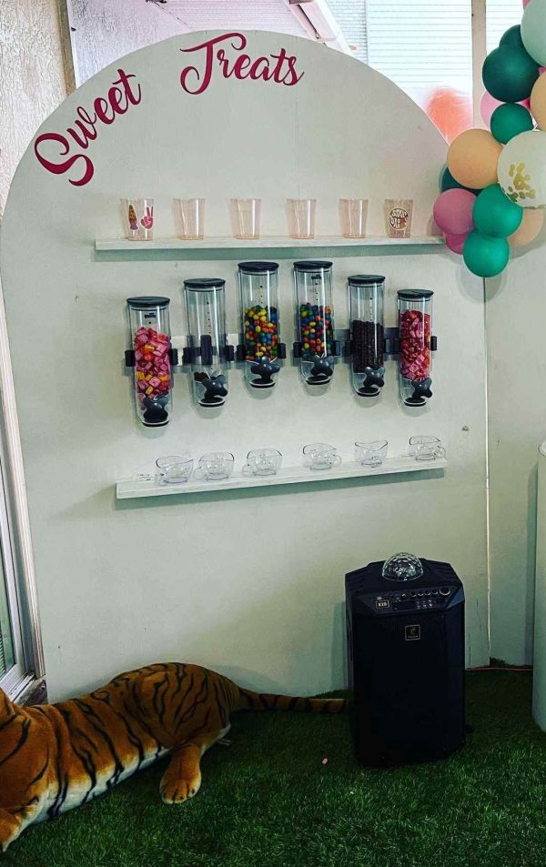 A candy stand labeled "sweet treats" with multiple jars of colorful candies, flanked by a Balloon Garland and a toy tiger on artificial grass.