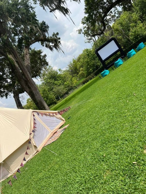 A canvas tent pitched on a grassy lawn in Orlando, with a movie screen and blue chairs set up in the background under hanging moss from trees.