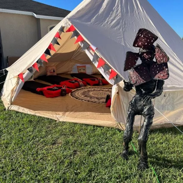 A person in a reflective polygonal suit stands in front of a large canvas tent decorated with bunting for outdoor glamping, set up on a grassy lawn.