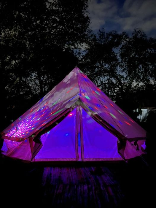 Backyard Glamping in Florida a Bell Tent illuminated by colorful lights against a dark, twilight sky, set in a wooded area for the ultimate sleepover experience.