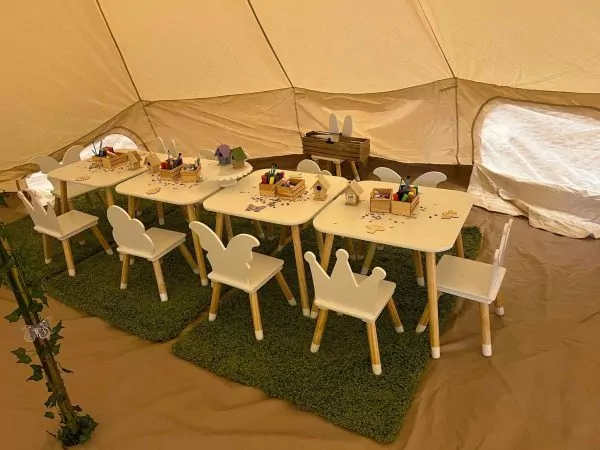 A children's activity area with small tables and chairs, crayons, and paper on a green carpet inside a tent for an outdoor birthday celebration.