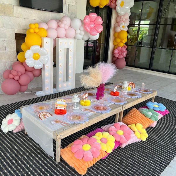 A colorful birthday party setup with balloon and flower-themed party themes decoration, a table laid with party favors, and lighted "one" sign, outdoors by a fireplace.