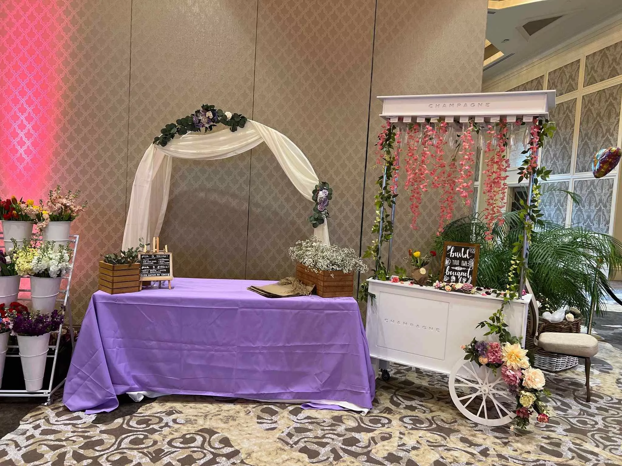 A wedding setup featuring a white arch with floral decorations, a draped table, and a decorative cart with flowers and balloons in an elegant hall for a Central Florida Corporate Event.