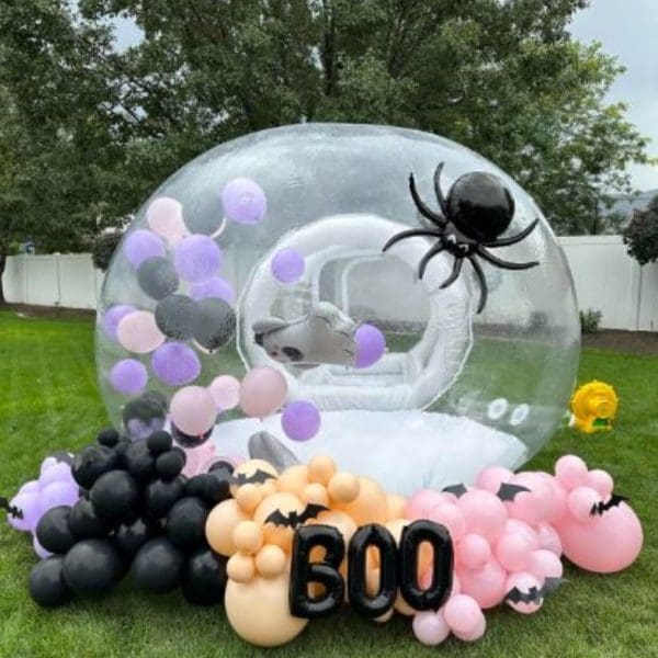 An Enchanting Bubble Balloon House Rental decorated with a black spider and balloons, surrounded by black and pink balloon clusters and a "boo" sign in a grassy yard.