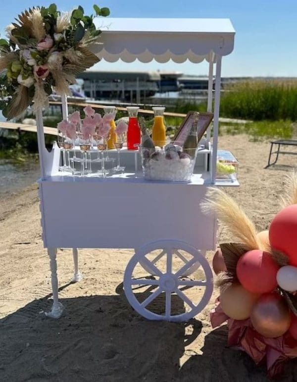 A small Elegant White Snack or Candy Cart Rental on a sandy beach decorated with flowers, offering drinks and snacks under a sunny sky.