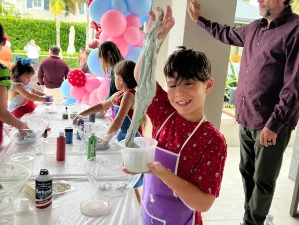 A young boy in a purple apron smiles while lifting a stretchy slime mixture at a crafts table during an outdoor kids party in Central Florida, with balloons in the background.