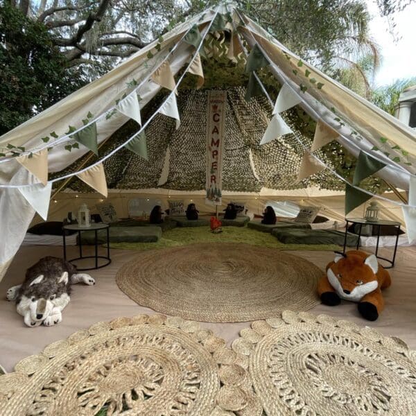 Step into the enchanting world of a cozy camping-themed paradise with this detailed interior view of a tent. The scene is adorned with lush leafy drapes and vibrant bunting, creating a whimsical atmosphere that invites adventure. Beneath your feet, round woven rugs offer a touch of rustic charm, while plush toy animals and glowing lanterns rest atop inviting green cushions. This captivating setup artfully combines elements of nature and comfort, making it the perfect haven for any celebration or playful escape. Ideal for those seeking camping party inspiration or cozy tent decor ideas!.