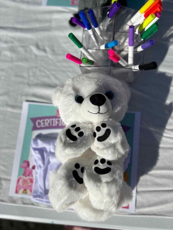 White teddy bear surrounded by colorful markers on a table with a certificate and a drawing displayed, ready for the 2024 Ultimate Sleepover.