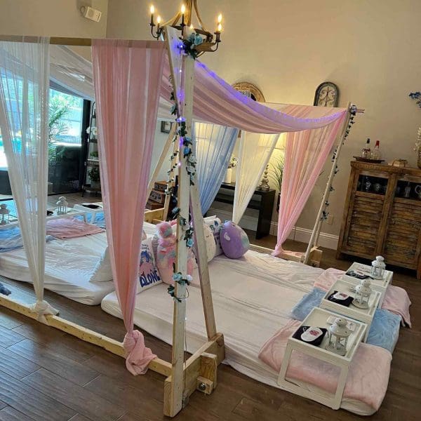 A cozy indoor canopy bed adorned with pink and white curtains, draped lights, and plush pillows, with a serving tray holding beverages beside it for the ultimate slumber party.