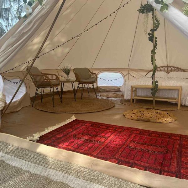 A cozy, furnished bell tent with chairs, a bed, and decorative rugs, set up outdoors under a tree for the ultimate night.