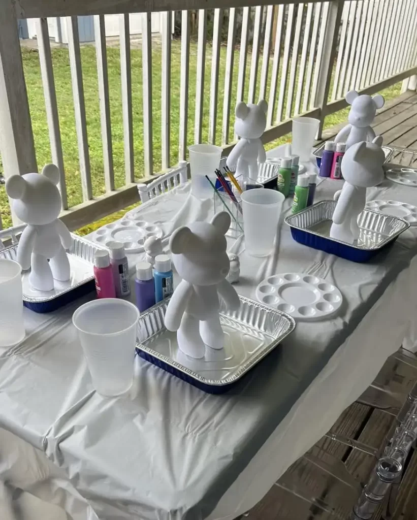 Barbie Themed Party white figurines on trays set up for painting, with brushes, colors, and cups on a covered table outdoors.