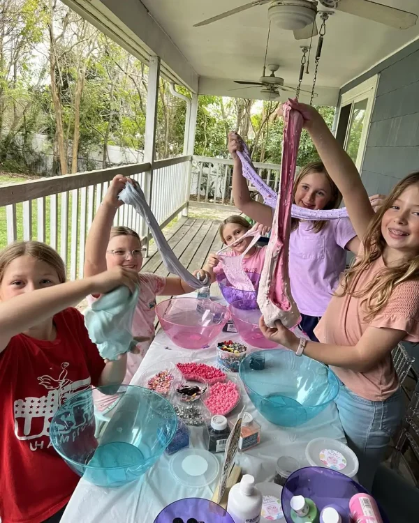 Four children making slime on a porch at a Barbie Themed Party, holding up their creations, with bowls and craft supplies on the table.