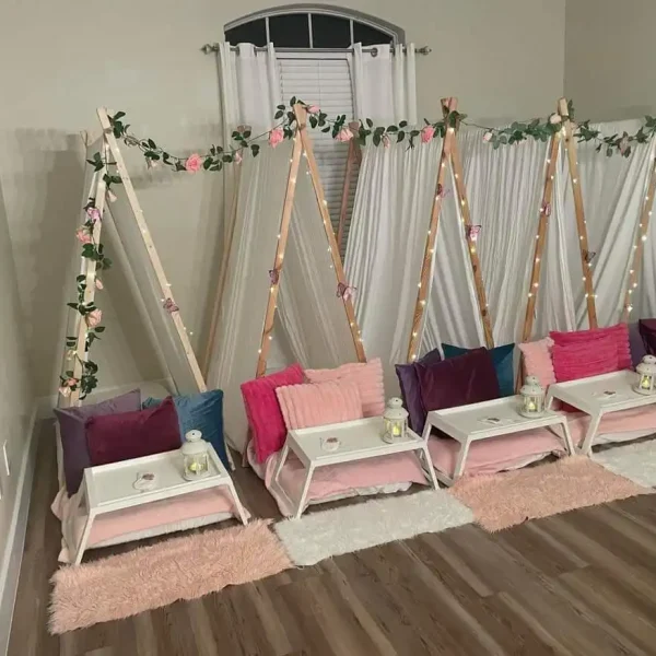 Indoor Barbie Themed Party setup with wooden teepees adorned with fairy lights and floral decorations, lined with colorful cushions and cozy rugs on a hardwood floor.