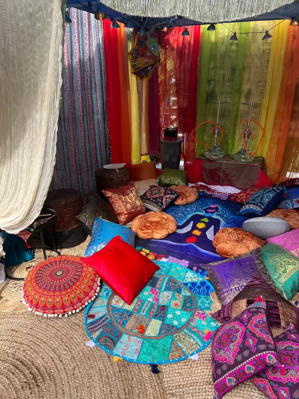 An elaborately decorated tent interior with colorful cushions, rugs, and hanging textiles, creating a vibrant Wonderland-inspired bohemian setting.