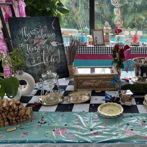 Elegant Alice in Wonderland party setup with a "there's always time for tea" sign, decorated tables with pastries and teapots, and a tropical backdrop.