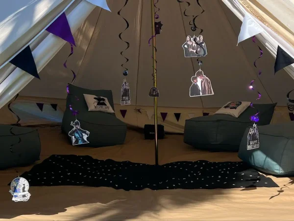 Interior of a tent decorated with a Wednesday Addams Party theme, including themed cushions, strings of lights, and purple and black accents.