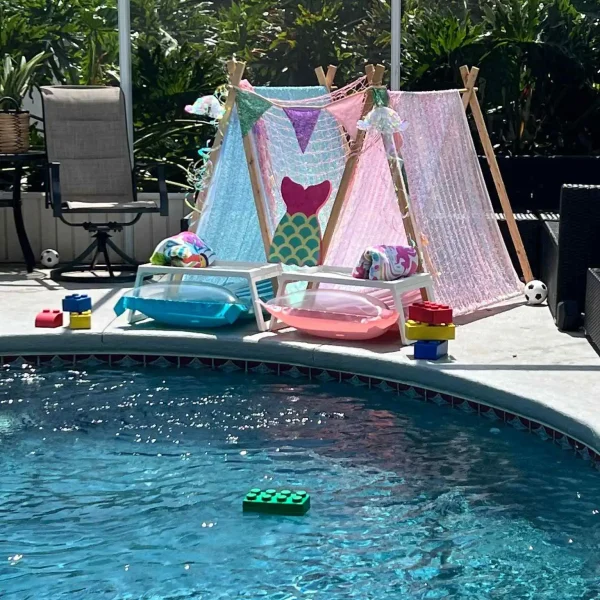 Transform your poolside experience with this enchanting setup featuring two stunning Teepee Cabanas beautifully adorned in vibrant mermaid-themed fabrics and colorful bunting flags. These whimsical cabanas set the scene for a perfect summer day. In front, find two cozy mini lounge chairs embellished with plush blue and pink cushions, ideal for relaxation under the sun. The pool area is playfully accentuated with large toy bricks lining the edge, some floating on the serene water surface, adding a touch of fun and creativity to the ambiance. All of this is nestled within a backdrop of lush greenery, creating a picturesque oasis that embodies tranquility and adventure—perfect for both kids and adults looking to unwind or play in style by the pool.