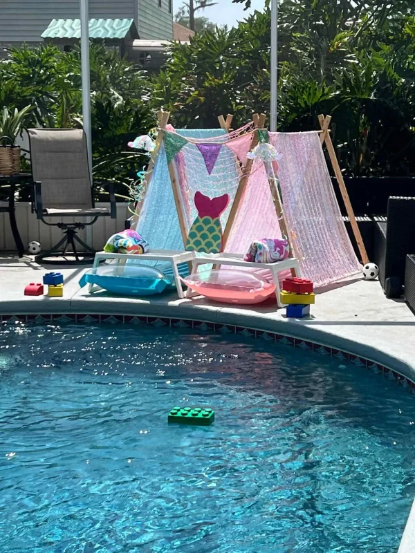 Transform your poolside experience with this enchanting setup featuring two stunning Teepee Cabanas beautifully adorned in vibrant mermaid-themed fabrics and colorful bunting flags. These whimsical cabanas set the scene for a perfect summer day. In front, find two cozy mini lounge chairs embellished with plush blue and pink cushions, ideal for relaxation under the sun. The pool area is playfully accentuated with large toy bricks lining the edge, some floating on the serene water surface, adding a touch of fun and creativity to the ambiance. All of this is nestled within a backdrop of lush greenery, creating a picturesque oasis that embodies tranquility and adventure—perfect for both kids and adults looking to unwind or play in style by the pool.