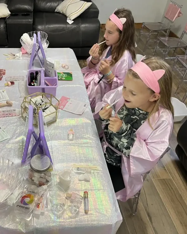 Two girls in pink headbands and robes applying makeup at a table filled with Spa Party with DIY lip Gloss during a Spa Party.