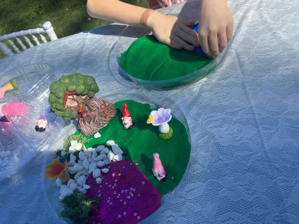 A young child creatively molds vibrant blue Play-Doh on a colorful plate, using green clay from the popular Play-Doh Sensory Table set for an imaginative play experience. Beside this scene, another plate artfully displays a miniature garden landscape complete with a miniature tree, tiny figurines, pristine white pebbles, and a delicate flower, capturing the joy of childhood creativity. This enchanting artistic display rests on an elegant white lace tablecloth illuminated by soft sunlight, enhancing its intricate details and inviting inspiration for imaginative playtime activities.