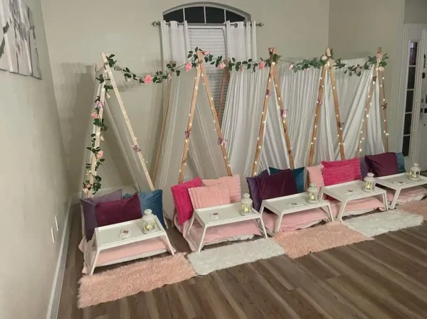 Indoor sleepover setup with wooden teepees, adorned with fairy lights and flowers for a Festival of Colors Party theme, lined with colorful cushions and small bedside tables.