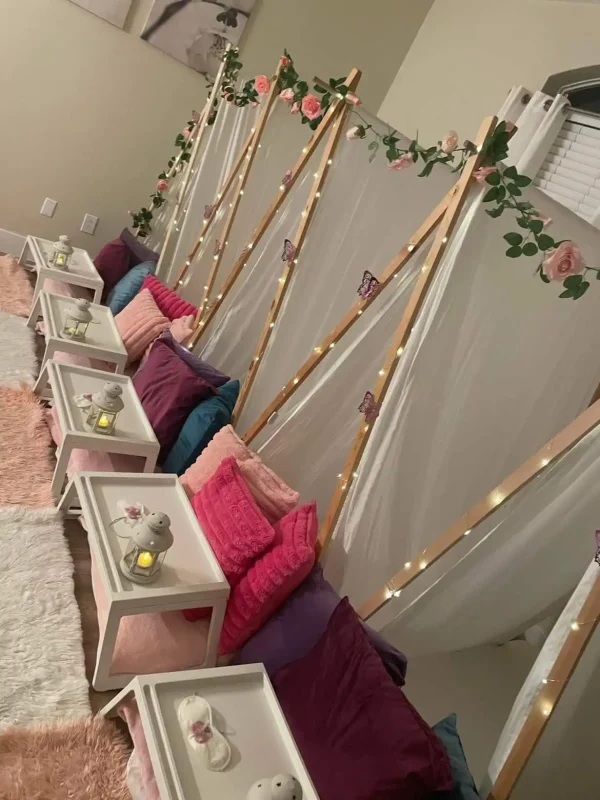 Row of cozy floor seating with cushions and blankets for a Dream Catcher Themed Party, each station divided by wooden frames adorned with string lights and roses.