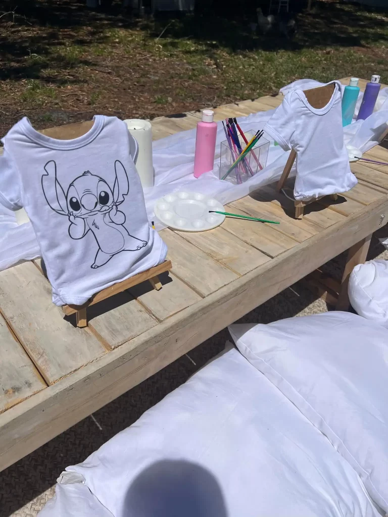 A white onesie featuring a printed cartoon character displayed on a wooden easel, surrounded by paint supplies and blank shirts on an outdoor table.