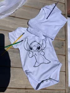 A white baby onesie with a creative twist, featuring a printed image of Disney's character Stitch on a wooden surface, alongside a yellow and a green pencil. Perfect for onesie painting parties at baby