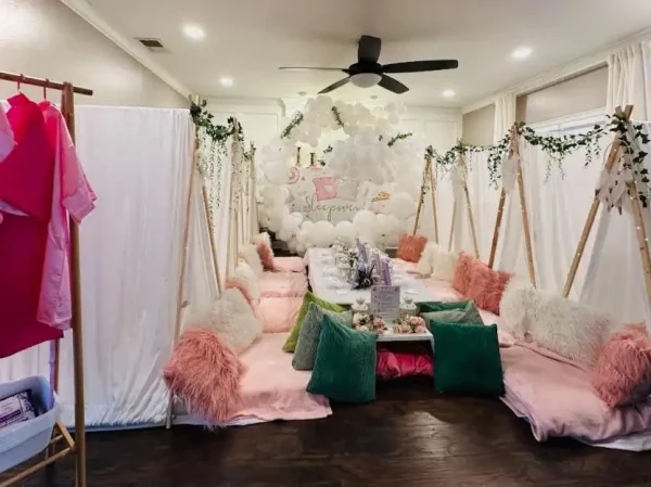 A room set up for a Spa Party with DIY Lip Gloss features low tables surrounded by cushions and faux fur blankets. Above the tables are white balloons, and foliage decorations hang from the ceiling. Add a touch of luxury with the DIY lip gloss station, perfect for an unforgettable slumber party experience.