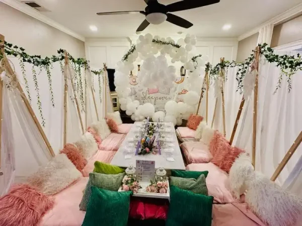 A cozy and inviting indoor dining setup features a low table surrounded by cushions and pillows, adorned with white balloons and greenery to create a serene atmosphere. A "Sleepover" sign in the background further enhances the theme. This setting is perfect for hosting a Spa Party with DIY lip Gloss, making it an ideal choice for a fun-filled time with friends!