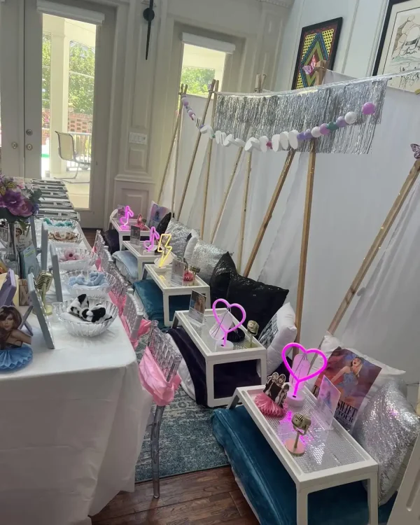 A room is set up for a Spa Party with DIY lip Gloss, featuring a row of small tents adorned with decorative pillows. In front of the tents, tables are arranged with light-up decorations, snacks, DIY lip gloss kits, and framed photos.