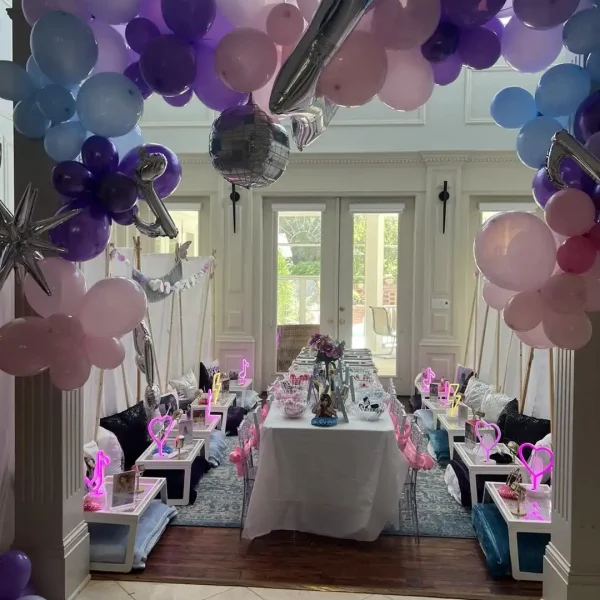 The "Spa Party with DIY Lip Gloss" transforms a room into a delightful children's spa setting with pink, purple, and blue balloons, a long dining table adorned with themed décor such as signs and miniature tents, and features an engaging DIY lip gloss station.