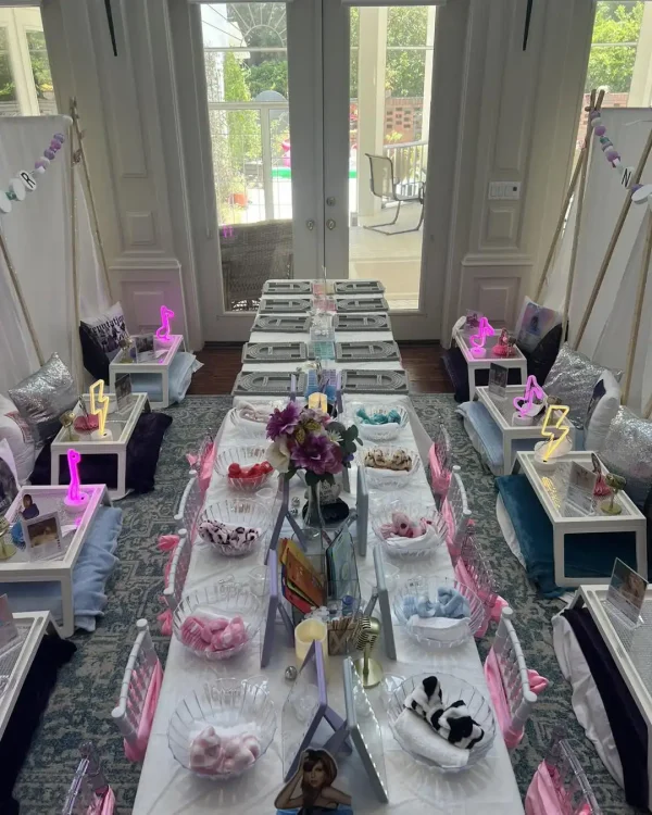 Create a slumber party setup with low tables, plates, cushions, and pink and white decor, featuring small tents and neon lights. Each place includes a tray with a photo, snacks, DIY lip gloss kits from the "Spa Party with DIY Lip Gloss," and a small pillow. Spa party vibes enhance the atmosphere as a pool shimmers in the background.