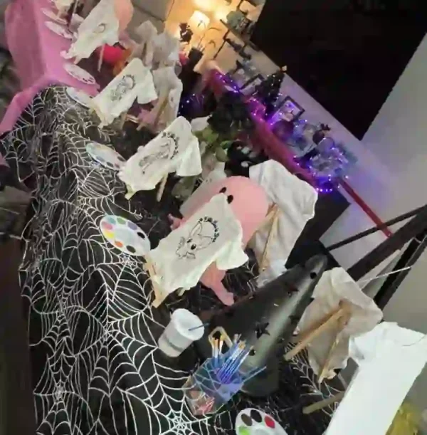 Engage in creative fun at your vibrant Crafty Onesie Painting Party baby shower! Picture the table, elegantly set with miniature easels showcasing canvases adorned with charming ghost designs, creating a delightful Halloween theme. A striking black tablecloth embellished with intricate white spiderweb patterns sets the tone for this festive occasion. Scattered across the table are colorful paint palettes, an assortment of brushes, and cups ready for artists to unleash their imagination. To complete this enchanting setting, soft purple string lights drape overhead, casting a cozy glow and enhancing the unique party atmosphere. Perfect for those seeking a spooky yet heartwarming celebration!