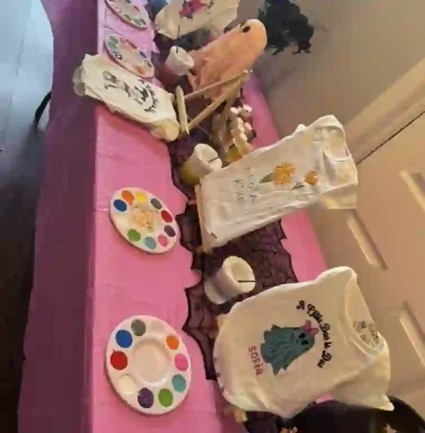 Get inspired for your baby shower with this vibrant Crafty Onesie Painting Party table setup! A chic pink tablecloth drapes elegantly over the table, serving as the perfect backdrop for an array of beautifully decorated onesies. Paint palettes and brushes are artfully arranged alongside a variety of crafting supplies, setting the stage for a creative and festive atmosphere. Whether you're planning a baby celebration or simply seeking unique decorating ideas, this detailed display is sure to captivate your guests and ignite their artistic flair.