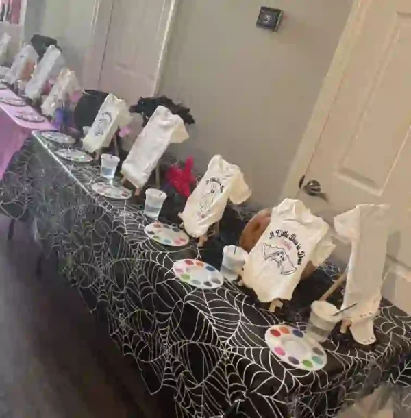 Elevate your baby shower with a stunning black-and-white spiderweb tablecloth, setting the stage for a memorable Crafty Onesie Painting Party! This elegantly themed setup features numerous easels showcasing white T-shirts, each adorned with intricate skeleton designs and playful phrases. Surrounding the display are vibrant paint palettes and ready-to-use cups of water, inviting guests to unleash their creativity in this artistic celebration. Perfect for an unforgettable DIY experience with friends and family!