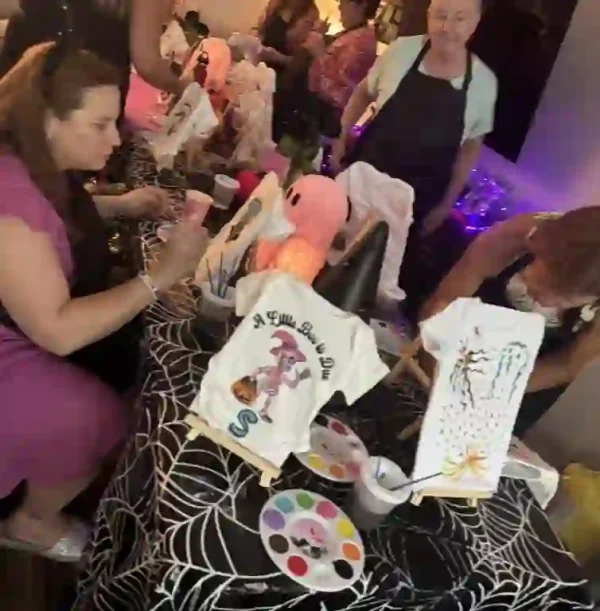 A lively group of friends gathered around a table during a Crafty Onesie Painting Party for an unforgettable baby shower experience. The table, adorned with a spooky black spiderweb-patterned tablecloth, serves as the creative hub where guests are enthusiastically painting vibrant designs on baby onesies and artful canvases. The dim ambiance is enhanced by twinkling string lights that cast a warm glow over the room, while a whimsical pink flamingo plush toy adds a touch of charm and playfulness to the celebration. This engaging scene perfectly captures the spirit of creativity and camaraderie at this unique baby shower event—ideal for expecting parents who love crafts and socializing in an inspiring atmosphere.