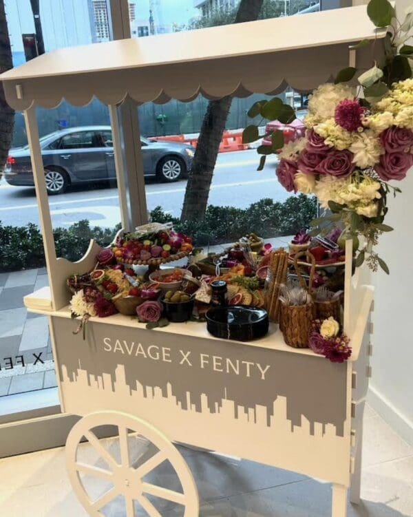 A stylish white cart adorned with the iconic "Savage X Fenty" branding displays an enticing assortment of vibrant fruits, exquisite flowers, and delectable snacks from the exclusive Movie Night Extravaganza collection. This eye-catching scene unfolds against a lively urban street backdrop, creating a captivating ambiance that perfectly blends luxury and casual elegance. Keywords: Savage X Fenty cart, Movie Night Extravaganza collection, vibrant fruits, exquisite flowers, delectable snacks, urban street scenery.